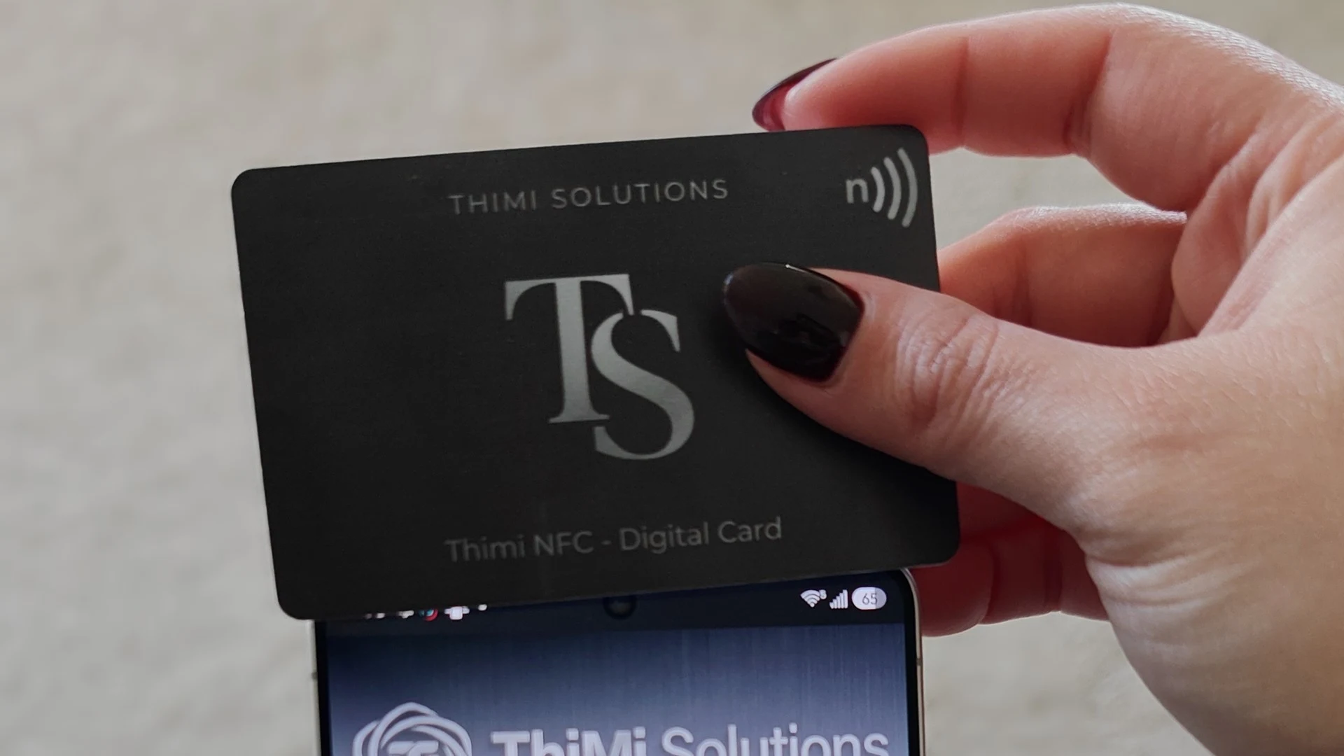 A black Thimi NFC card tapping the top of an Android phone, which opens a digital business card profile.